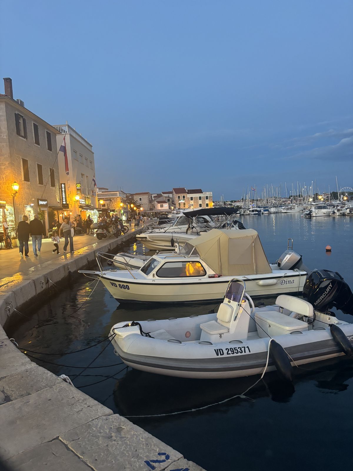 Boote am Kai in Vodice in der Dämmerung, dahinter die beleuchtete Uferpromenade und die Marina